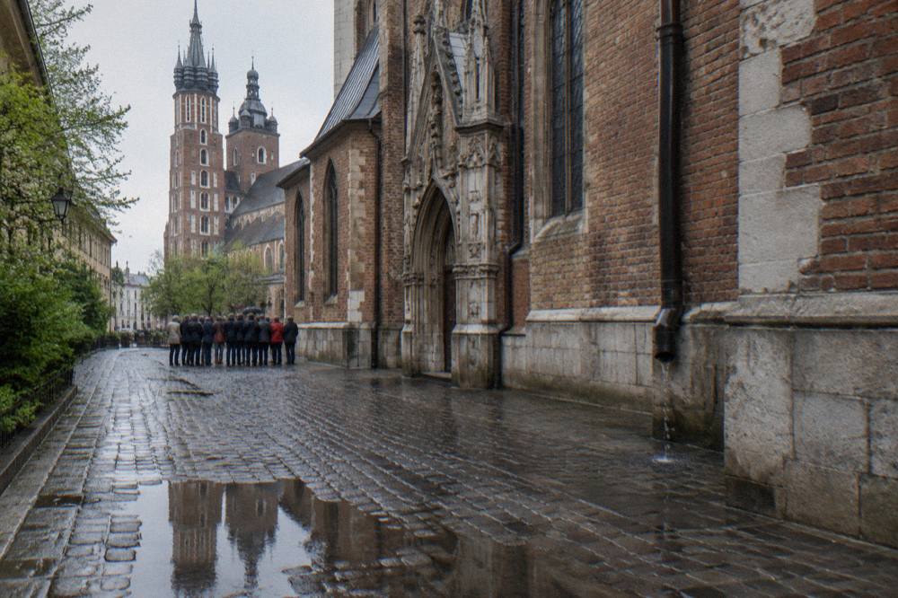A small group stands on wet cobbles beside a Gothic brick church, with Krakow church towers and a puddle reflection in the distance.