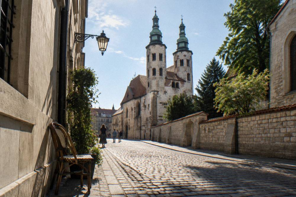 Cobblestone street in Krakow with St Andrew's Church towers ahead, stone walls, trees, a wall lamp, an empty chair, and distant walkers.