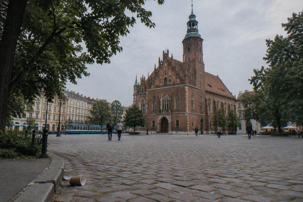Cobbled Wolnica Square with the brick Kazimierz Town Hall, leafy trees, distant people, a tram, and a tipped paper cup by the kerb.