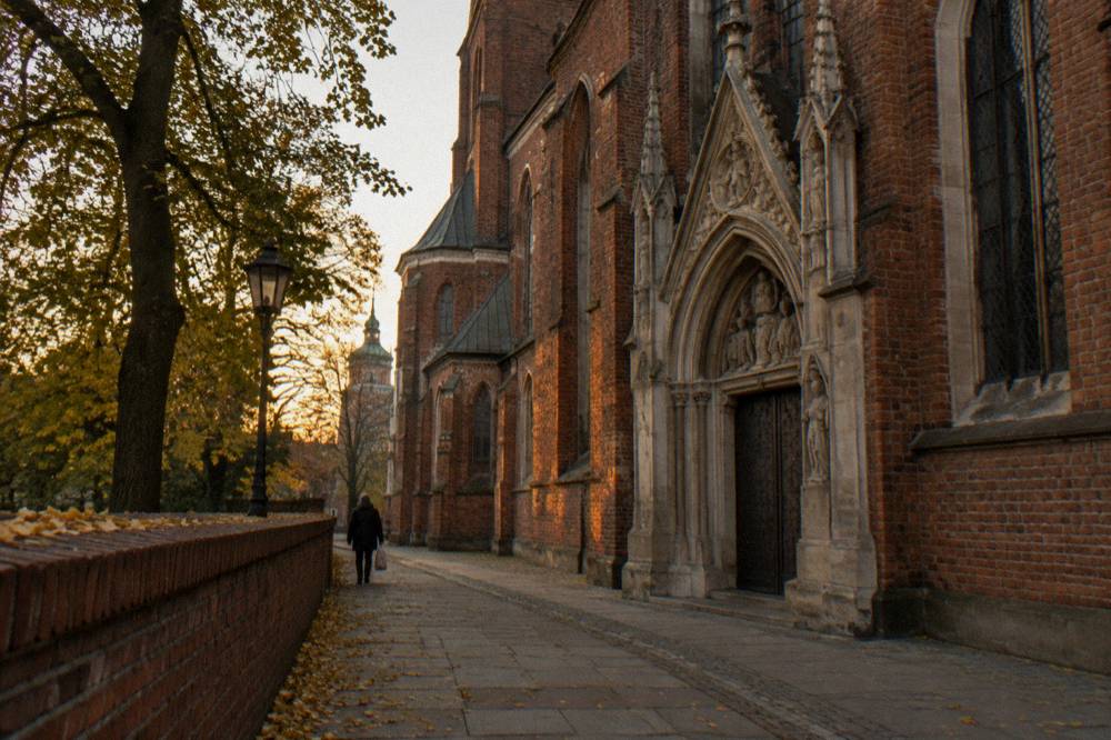 Red brick Gothic church wall and stone portal beside a tree-lined path with fallen leaves and a distant pedestrian at dusk.
