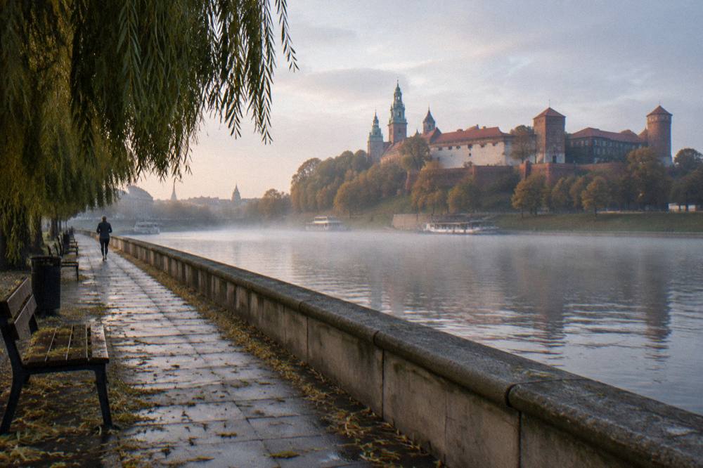 Wet riverside promenade beside the Vistula with a distant jogger, willow branches, mist on the water, and Wawel Castle on the hill beyond.