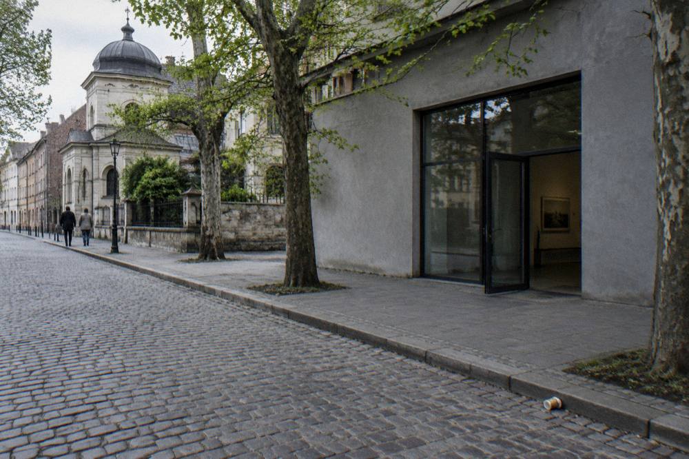 Entrada moderna de color gris del museo con una puerta de cristal abierta junto a una calle empedrada, plátanos de sombra y edificios históricos de Kazimierz.
