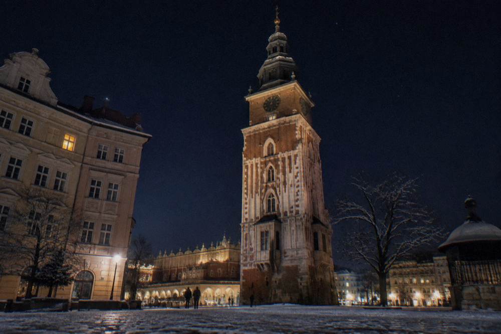 La Torre del Ayuntamiento de Cracovia iluminada se alza sobre una Plaza del Mercado nevada de noche, con el Lonja de los Paños y figuras borrosas cerca.