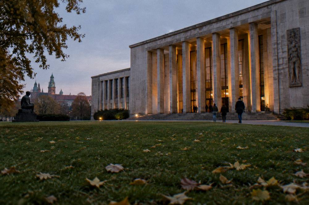 Fachada de museo modernista iluminada con altas columnas de piedra, césped con hojas caídas, visitantes lejanos en la entrada y las torres de Wawel al fondo.