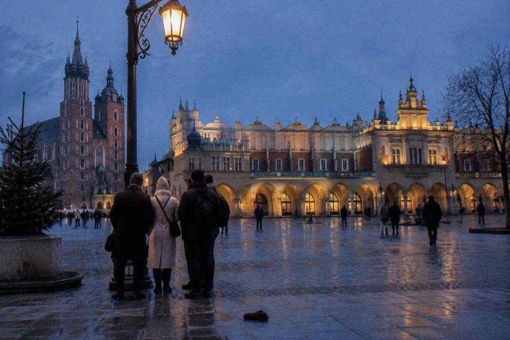 Un grupo pequeño bajo una farola encendida en el pavimento mojado de la Plaza del Mercado de Cracovia al anochecer.