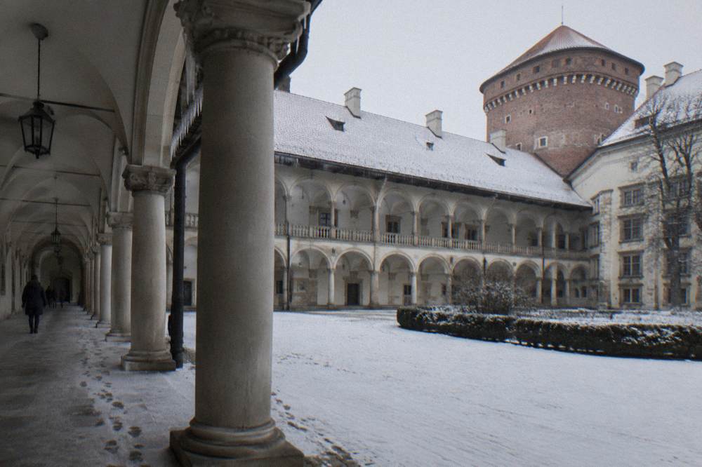 Snow-dusted Wawel Castle courtyard with pale arcades, columns, footprints, bare trees and a brick tower under a gray winter sky.