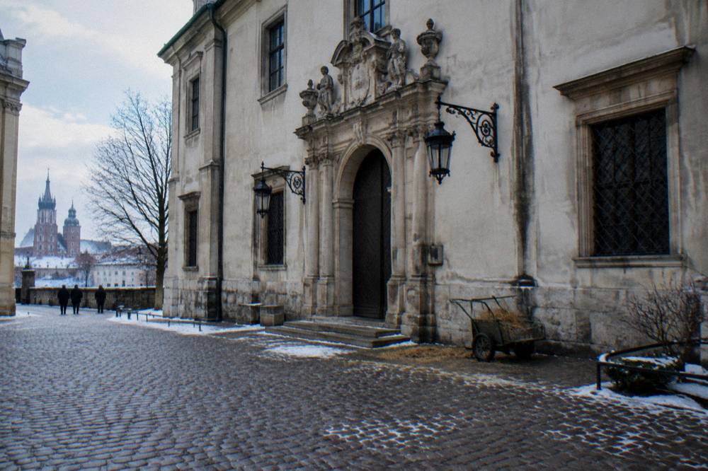 Snow-dusted cobbles lead past a baroque stone portal with iron lanterns and a trolley, with Krakow church towers in the distance.