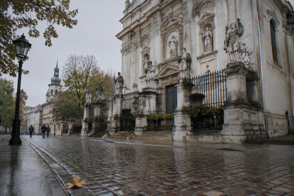 Wet cobbles lead past an iron fence and stone apostle statues in front of a Baroque church facade on a tree-lined Krakow street.