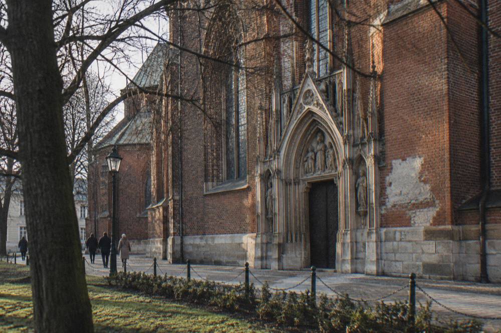 Bare trees frame a red brick Gothic church wall with a carved stone portal and a few pedestrians on a winter path.