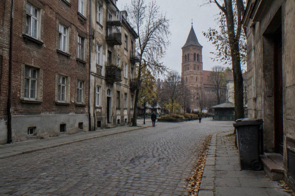 Calle empedrada tranquila con edificios bajos de ladrillo, un contenedor con ruedas, árboles sin hojas y una torre de iglesia bajo un cielo gris.