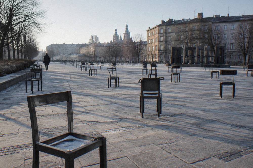 Filas de sillas de metal vacías sobre una plaza empedrada con escarcha, árboles sin hojas, bloques de apartamentos antiguos y una persona a lo lejos.