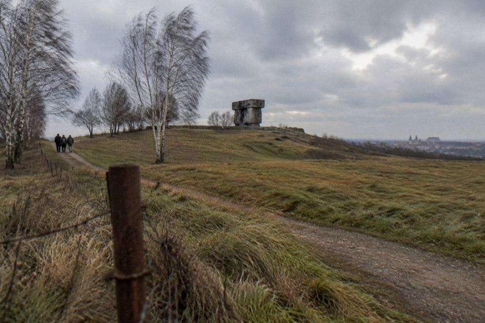 Colina ventosa con hierba, abedules desnudos, un camino de tierra, un poste oxidado y un gran monumento de piedra bajo un cielo nublado