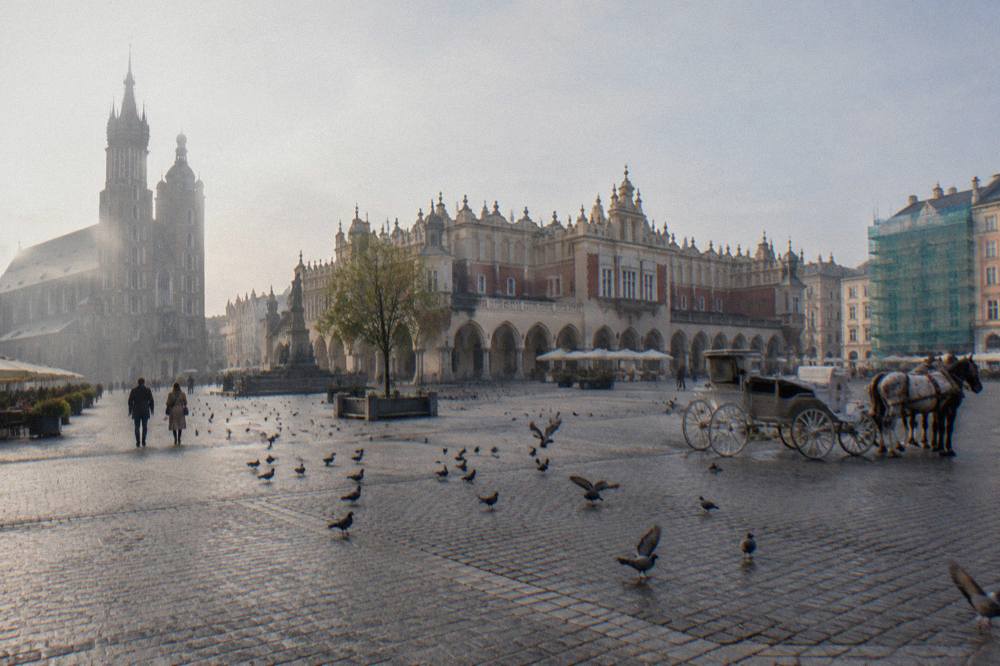 Plaza del Mercado de Cracovia empedrada, con el Pabellón de los Paños, la basílica de Santa María, palomas, un coche de caballos y casas con andamios.