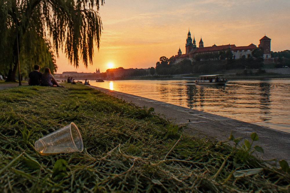 Un vaso de plástico tirado en la hierba segada junto al Vístula al atardecer, con sauces, gente a lo lejos, un barco y el castillo de Wawel al fondo.
