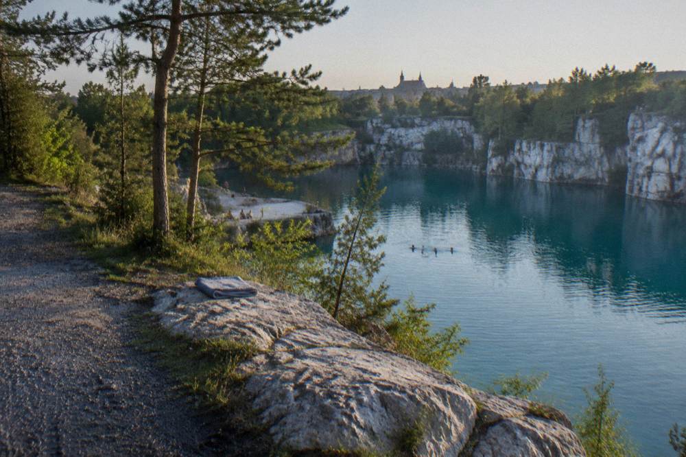 Lago de cantera turquesa bajo acantilados de caliza clara, pinos, un camino de grava y una toalla doblada sobre un saliente rocoso.