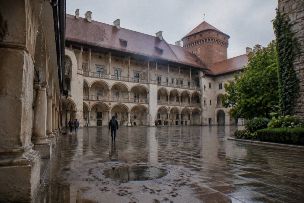 Patio de las Arcadas del Castillo de Wawel mojado con losas reflectantes, una torre de ladrillo, árboles y algunas personas a lo lejos tras la lluvia.