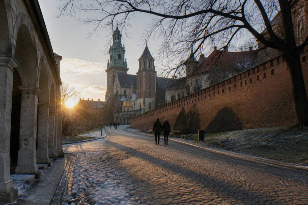 El sol bajo de invierno ilumina un camino empedrado junto a los muros de ladrillo de Wawel, galerías, árboles sin hojas, parches de nieve y paseantes a lo lejos.