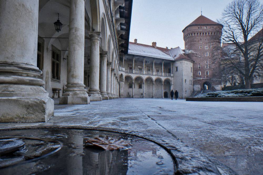 Patio del Castillo de Wawel con galería de arcos, torre redonda de ladrillo, árboles sin hojas y una hoja congelada en un charco poco profundo.