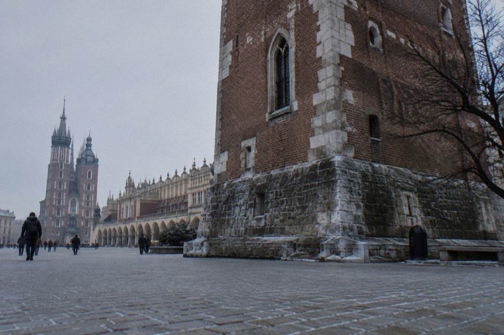 Vista invernal baja de la Plaza del Mercado de Cracovia con la base de la Torre del Ayuntamiento, el Lonja de los Paños, la basílica de Santa María y peatones a lo lejos.