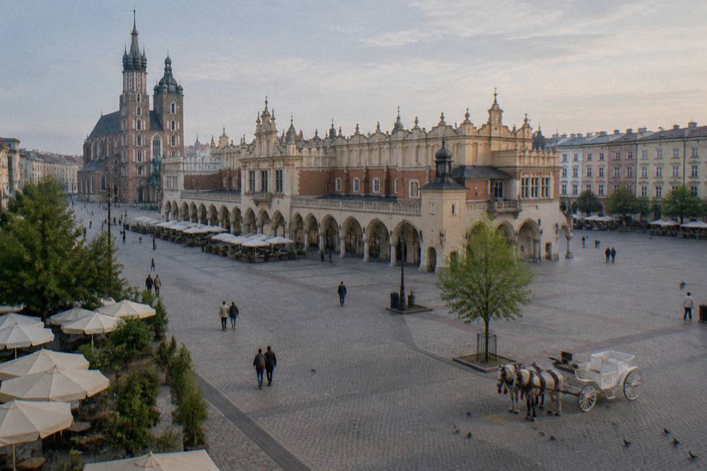 Vista elevada de la Plaza del Mercado de Cracovia con la Lonja, la basílica de Santa María, fachadas pastel, peatones y un carruaje tirado por caballos.