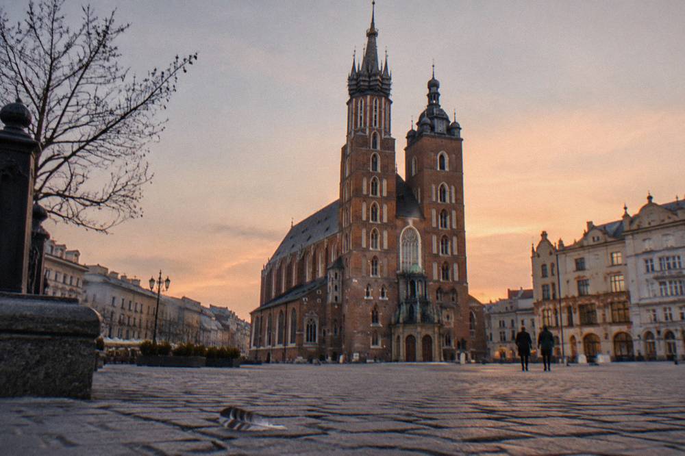La basílica de Santa María se alza sobre el empedrado de la plaza del mercado de Cracovia al atardecer, con ramas desnudas, peatones lejanos y una pluma en primer plano.