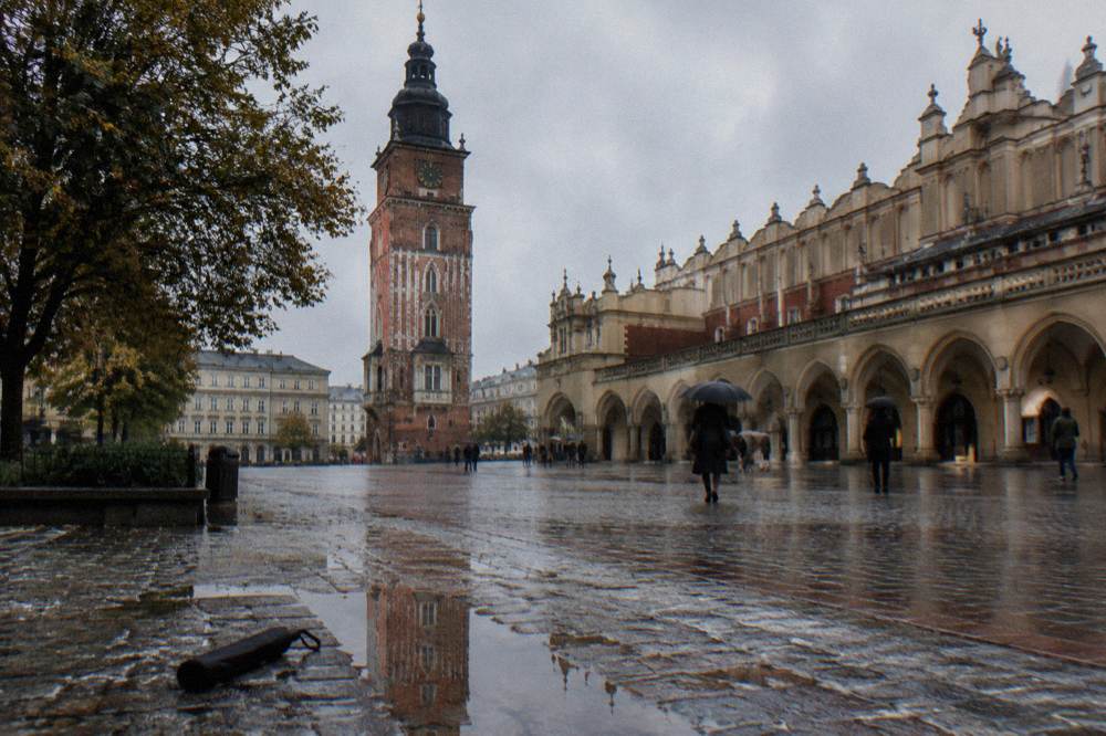 Plaza del Mercado con adoquines mojados, la Torre del Ayuntamiento, las galerías de la Lonja de los Paños, paraguas, árboles y un charco que refleja el entorno.
