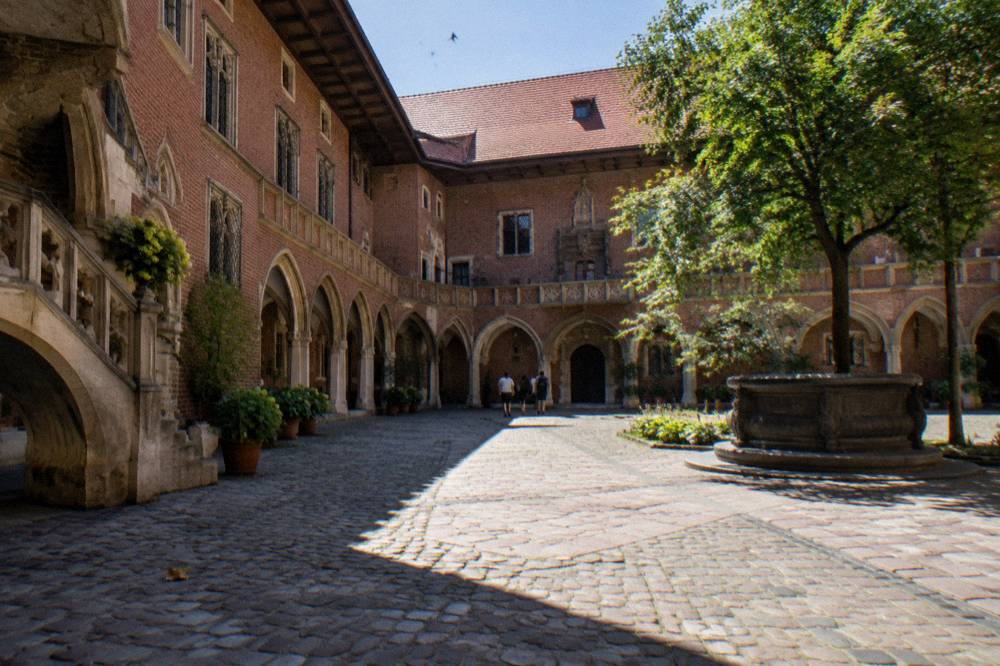 Patio gótico de ladrillo iluminado por el sol, con arcadas de piedra, adoquines, un árbol, un pozo y algunas personas a lo lejos.