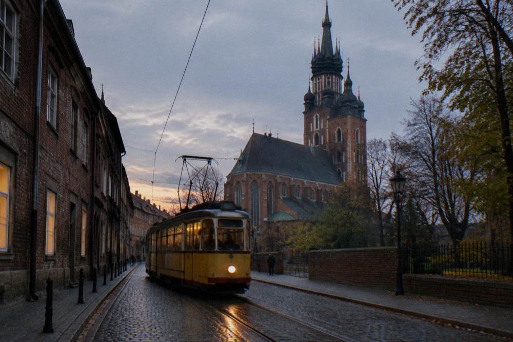 Un tranvía amarillo antiguo circula por adoquines mojados junto a edificios de ladrillo, con una torre gótica de iglesia elevándose tras los árboles al atardecer.