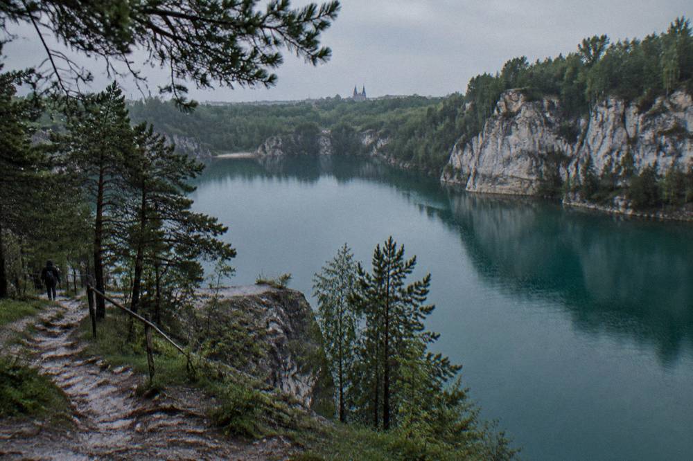 Laguna de cantera turquesa bajo acantilados de caliza y pinos, con un estrecho sendero y un caminante lejano bajo un cielo gris.