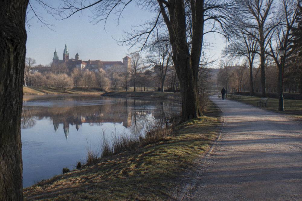 Un camino de grava discurre junto a aguas quietas y árboles desnudos, con un paseante lejano y el Castillo de Wawel al otro lado del agua.