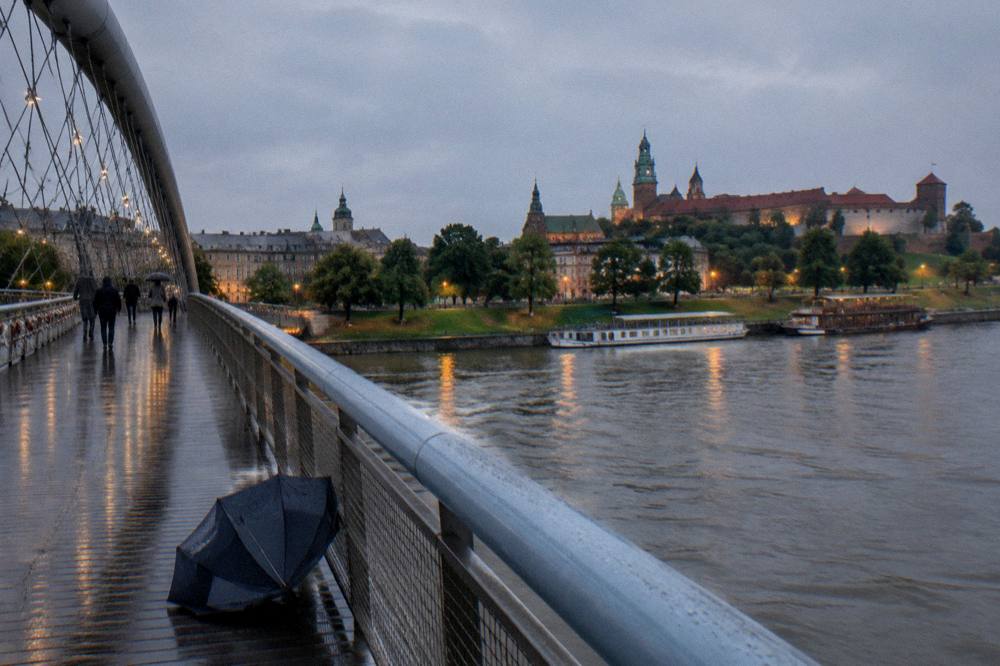 Pasarela del puente peatonal sobre el Vístula con un paraguas abandonado, peatones lejanos, barcos fluviales y el Castillo de Wawel al otro lado del río.
