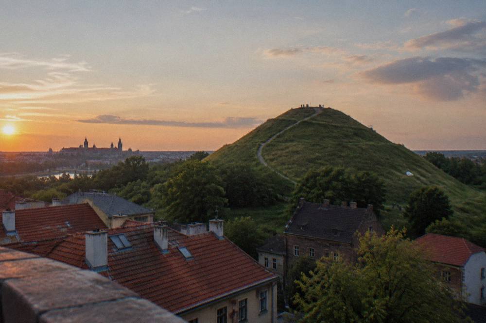 Un túmulo cubierto de hierba con un camino gastado se alza sobre los tejados de teja roja al atardecer, con las torres lejanas de Cracovia en el horizonte.