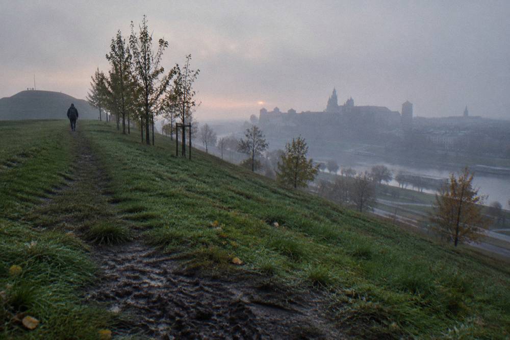 Un camino de hierba con rocío asciende por un túmulo arbolado al amanecer brumoso, con un único paseante y el horizonte de Cracovia apenas visible al otro lado del Vístula.