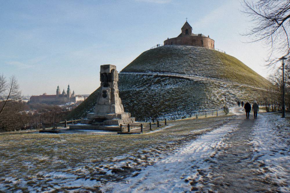 El túmulo de Kościuszko, cubierto de nieve, se alza tras un monumento de piedra, con una capilla de ladrillo en la cima y personas caminando por un sendero embarrado.