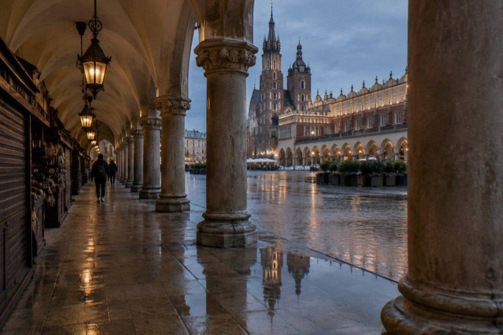 Wet stone arcade beside shuttered stalls, with columns, lamps, reflections, and St Mary's Basilica across Krakow's market square.