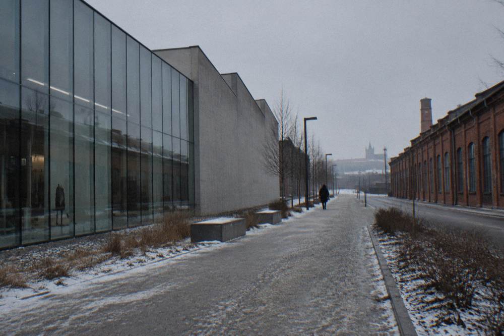 Snow-dusted walkway between a glass museum facade and red brick factory buildings with a lone person in the distance.