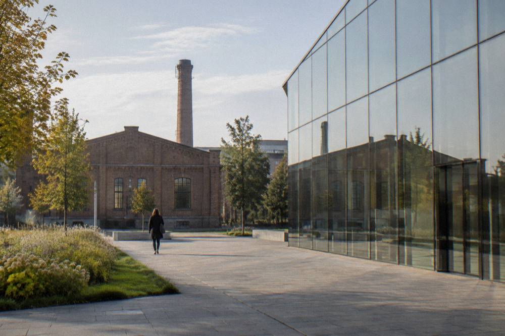 A lone person walks across a paved museum forecourt beside a glass facade, with brick factory buildings and a chimney behind.