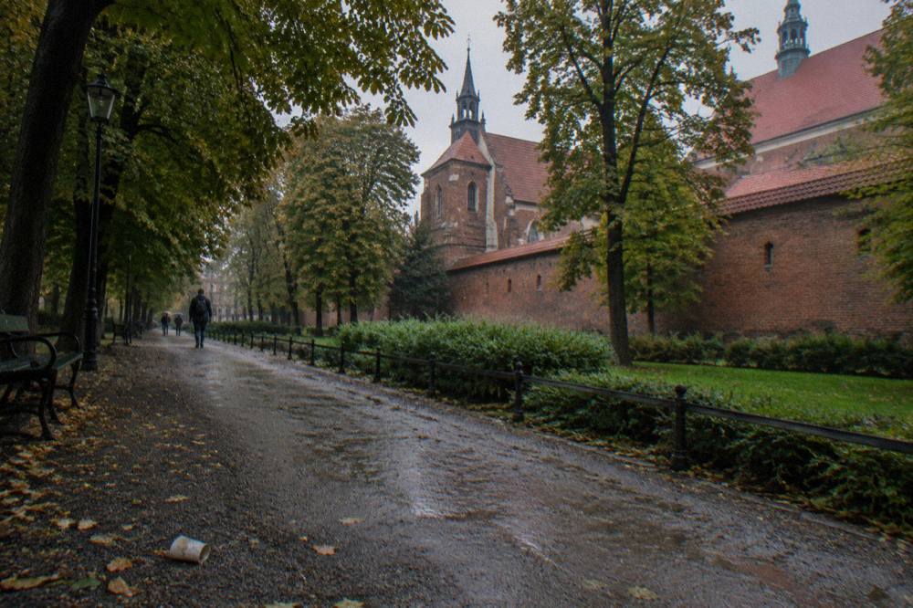 Wet tree-lined park path with benches, lamp posts, low railings, distant walkers, and a red-brick church wall.