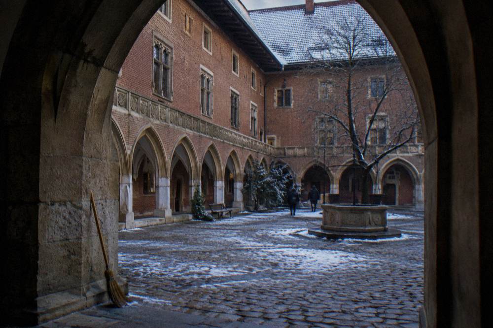 Snow-dusted cobbled university courtyard with brick Gothic arcades, a stone well, bare tree, and broom seen through a dark archway.