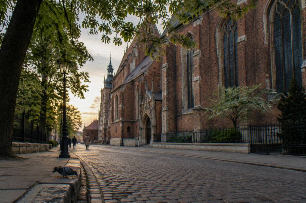 A red brick Gothic church lines a cobbled street with leafy trees, a lamppost, distant pedestrians and a pigeon on the kerb.