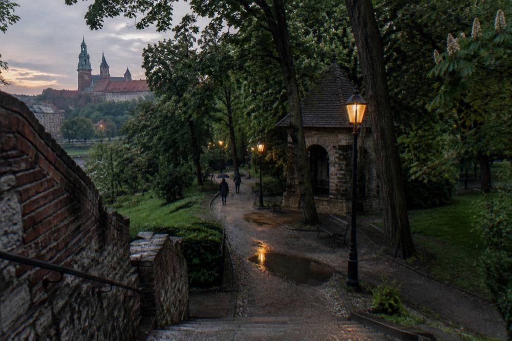 A wet park path descends past stone walls, lamps, trees and a small stone shelter, with castle towers in the distance.