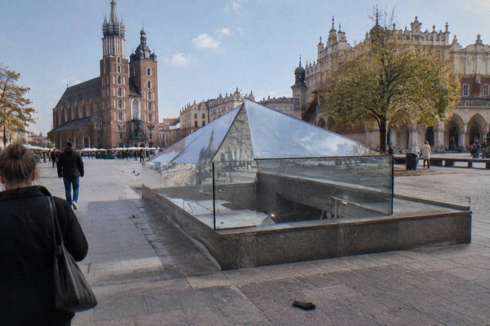 Entrada de cristal del museo subterráneo en la Plaza del Mercado de Cracovia, con la basílica de Santa María y la Lonja de los Paños al fondo.