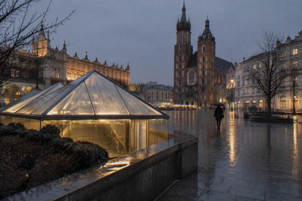 Techo de cristal del pabellón del Rynek Subterráneo brilla sobre los adoquines mojados de la plaza del mercado con la basílica de Santa María al fondo.