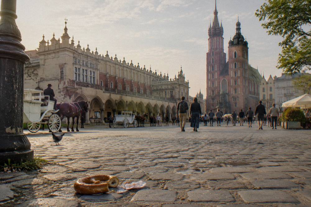 Plaza del mercado empedrada de Cracovia con las galerías del Sukiennice, las torres de la basílica de Santa María, coches de caballos, peatones y un pretzel.