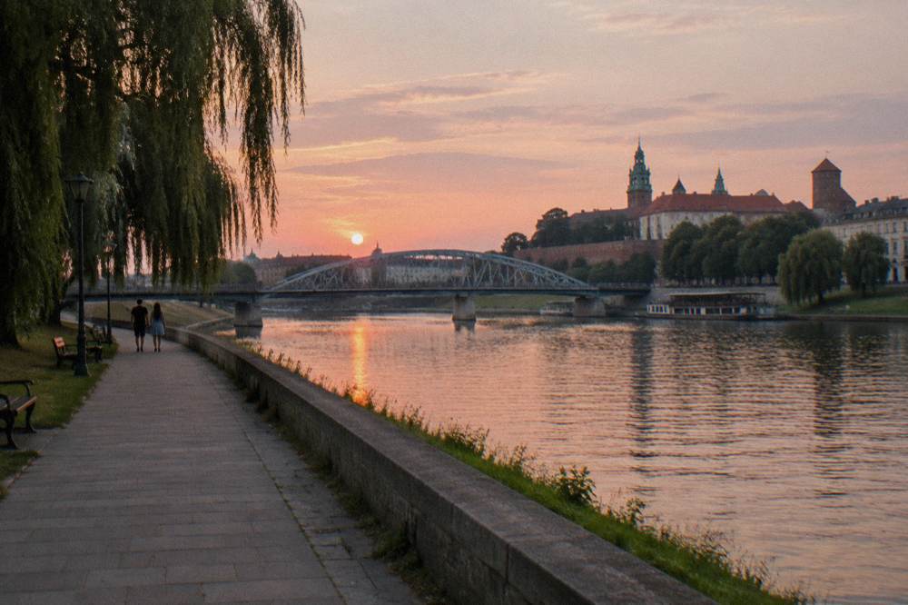 Dos paseantes lejanos avanzan por el paseo fluvial del Vístula al atardecer, con el Castillo de Wawel, un puente de acero y sauces al otro lado del río.