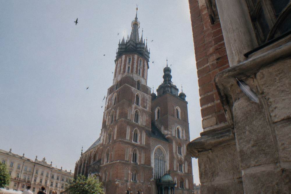 Las torres de la basílica de Santa María se alzan sobre los tejados de Cracovia bajo un cielo pálido, con pájaros y una pluma atrapada en un saliente de piedra.