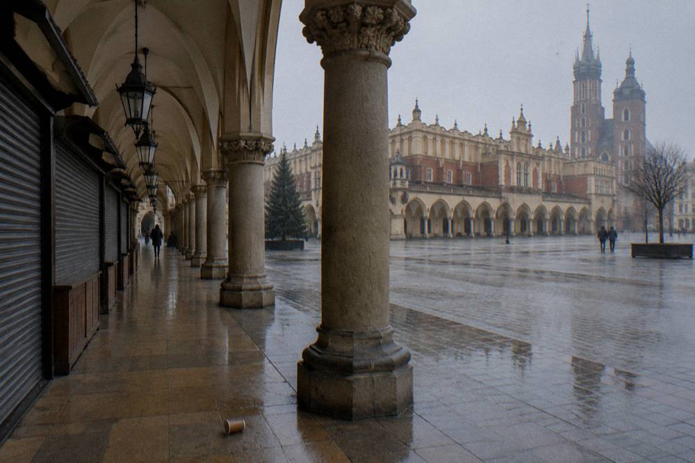 Arcada del Sukiennice mojada, con puestos cerrados, columnas de piedra, un vaso de papel en el suelo y la basílica de Santa María al otro lado de la plaza.