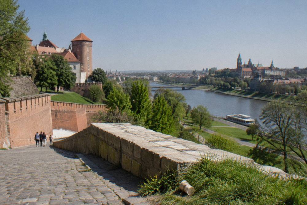 Cobbled Wawel Hill slope with brick castle walls, a stone parapet, grass, distant people and the Vistula River under a clear sky.