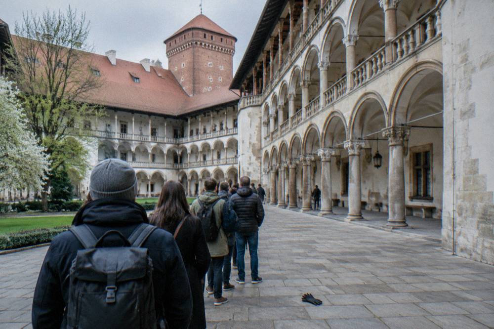 Visitors queue from behind in Wawel Castle's arcaded courtyard, with a brick tower, stone paving, spring trees and a dropped glove.