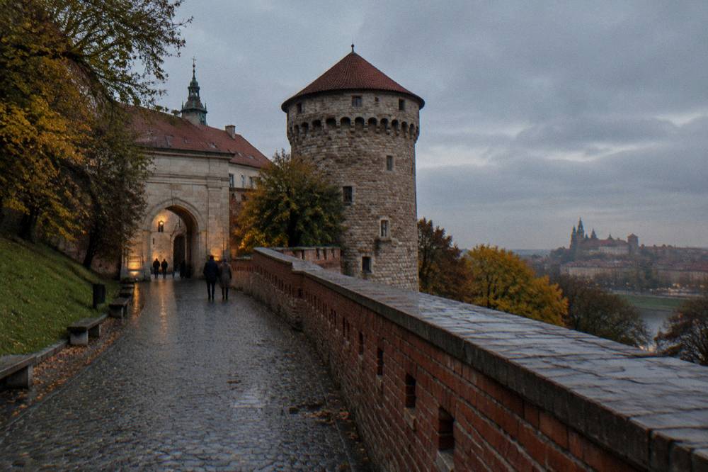 Wet cobbled path and brick defensive wall lead to a round stone tower and arched gateway on Wawel Hill at dusk.
